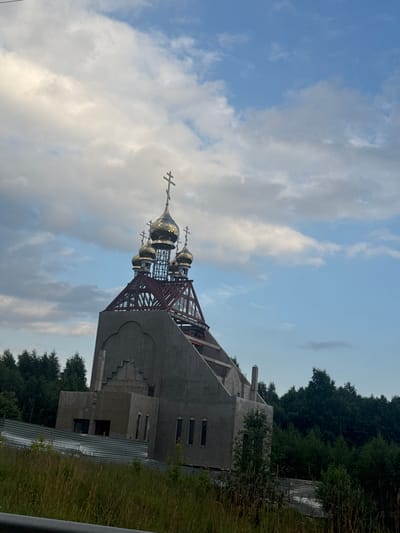 The image depicts an unfinished Orthodox church under construction in Novoe Devyatkino, Russia. The main structure is a concrete building, with a section of the roof open to reveal a metal framework. Topped with golden domes and crosses, this structure suggests a prominent religious building. The scene is set against a backdrop of lush green trees, likely a forest, and a sky filled with scattered clouds. The time of day appears to be late afternoon or early evening, as indicated by the lighting and the soft hues of the sky. The overall atmosphere is one of ongoing development and anticipation, with the church not yet complete but already showing its distinctive architectural features. There is no visible text in the image.