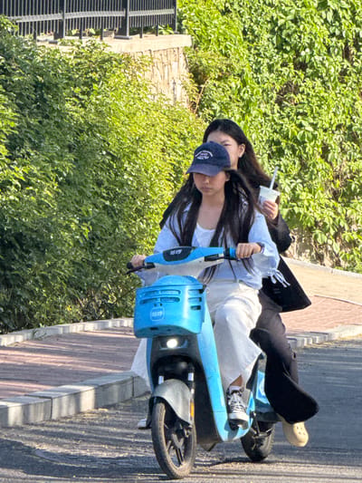 Two young women ride a blue electric scooter on a paved area adjacent to a hedge. The driver wears a baseball cap, white shirt, and white pants, while the passenger behind her wears a black jacket and pants. The location appears to be Zhongtun, China.