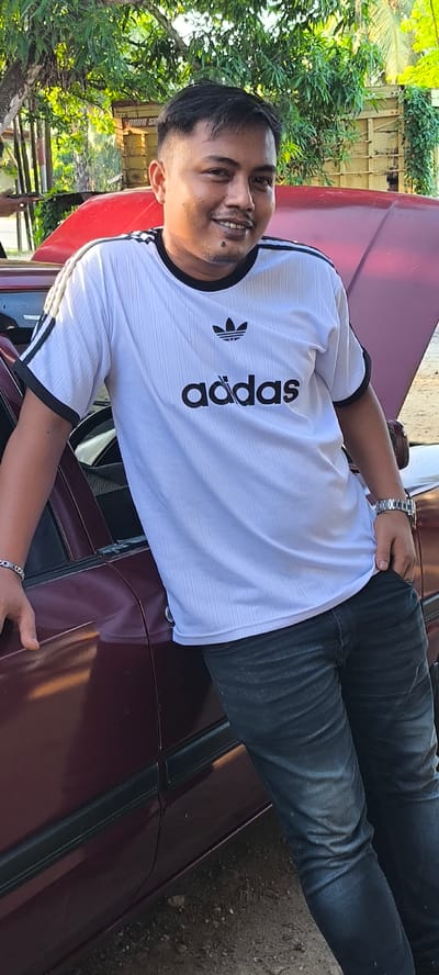 A full-body shot shows a man with short, dark hair and a light complexion leaning against a dark red car with its hood open. The man is wearing a white t-shirt with black stripes on the sleeves and collar, and the Adidas logo and name printed in black on the front. He is also wearing dark wash jeans. His left arm is resting on the car door, and his right hand is in his pocket. He is smiling at the camera. The car appears to be parked on a dirt road, with trees and some structures visible in the background. The scene is outdoors and appears to be in a rural or semi-rural area. The image is taken from a slightly low angle, with the man filling most of the frame. The overall impression is casual and relaxed. The location is Aluebutuha, Indonesia.