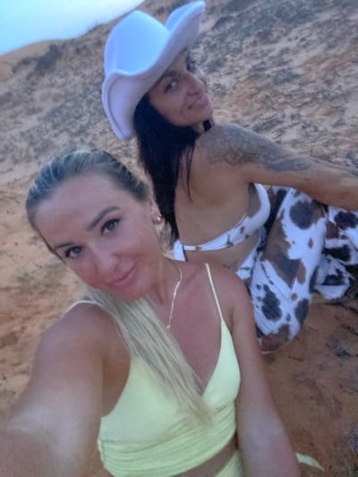 Two women are sitting in a sandy environment. The woman in the foreground, on the left, has blonde hair and is wearing a light yellow top. She is looking towards the camera, smiling. The woman in the background, on the right, is wearing a white cowboy hat and a bikini with a cow print pattern. She is also looking towards the camera and smiling. The background is a sandy hill. The scene appears to be outdoors, likely in a desert or beach-like setting. While the specific location of Mui Ne, Vietnam, is mentioned in the prompt, there are no visual cues in the image to confirm this.
