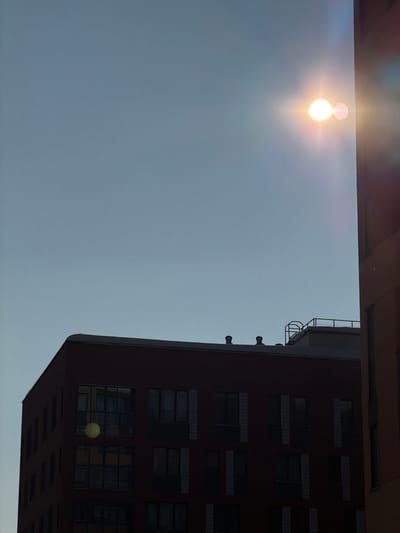 The scene depicts an urban landscape in Средний Постол, Russia, under a clear, light blue sky. The sun, a prominent bright white and yellow circular light source, is positioned in the upper right quadrant of the frame, emitting strong lens flare and a pinkish secondary flare artifact to its right.
In the lower left and central areas of the image, a multi-story building with dark reddish-brown exterior walls is visible, largely in shadow. This building features numerous rectangular windows arranged in a grid pattern; one window in the lower left reflects a yellowish circular lens flare artifact. The rooftop of this building displays two dark, circular ventilation structures.
To the right, a vertical section of a second, taller building with a lighter reddish-brown facade is partially visible. This building also appears to be in shadow and exhibits vertical architectural elements. Behind the roofline of the main reddish-brown building, a lighter colored structure with a metal railing or platform on its top is discernible, suggesting another building or rooftop installation further in the background. No individuals, overt actions, or interactions are present in the image.