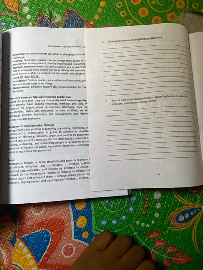 An open textbook, displaying academic content on "Basic Principles of Entrepreneurship" and detailing "Management and Leadership" concepts including leadership qualities and definitions, rests on a surface. The visible page, numbered 24, includes questions with blank lines for responses. The textbook is positioned on a colorful, embossed plastic mat featuring a repeated geometric and organic pattern in shades of green, blue, yellow, red, and white. A light-skinned human hand, with fingers slightly curled, is partially visible in the lower-left foreground, resting on the patterned mat. The scene suggests an indoor study or reading environment in Nsukka, Nigeria.