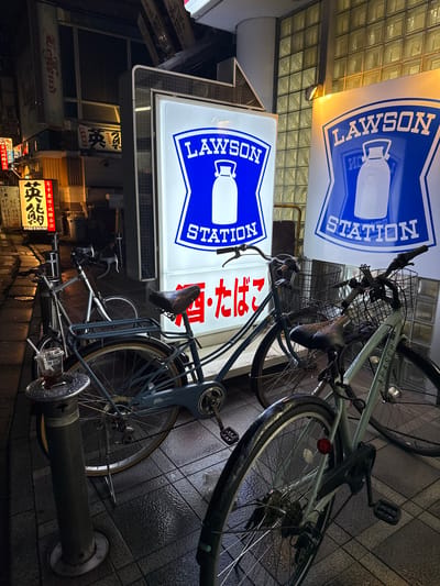 This media file is a nighttime, eye-level photograph taken outdoors in a Japanese urban setting, likely Adachi, Japan. The scene is illuminated by neon signs, typical of a streetscape in Japan. Two prominent blue and white "LAWSON STATION" signs are visible, with the text "LAWSON" and "STATION" in white capital letters against a blue background. One sign is positioned above the other, both displaying a milk bottle icon.
Several bicycles are parked and leaning against a wall or each other. One bicycle in the foreground is dark blue with silver accents, featuring a wire basket, disc brakes, and a red reflector. Another bicycle, a lighter color, is visible behind it. The ground is paved with square tiles, which appear wet, suggesting recent rain or a damp evening.
Various other signs in Japanese characters are also visible, some illuminated and some darker. One sign has the text "年中無休・明瞭会計" (Open all year round, clear accounting). Another sign reads "酒・たばこ" (Alcohol and tobacco). The overall atmosphere is that of a commercial street or an alleyway at night, with the glow of the signs casting light on the wet pavement and the parked bicycles. There are no people visible in the frame, and the image has a slightly tilted perspective.