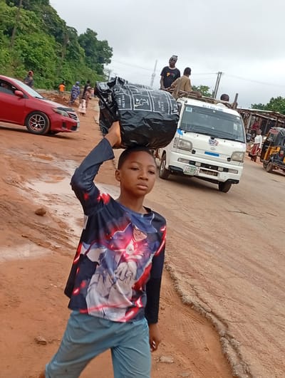 In Nkwelle Ezunaka, Nigeria, a young boy with a serious expression carries a large black bag on his head, steadying it with his hand. He wears a long-sleeved shirt with a graphic design and light blue pants. The road is unpaved, reddish-brown, and shows signs of disrepair. Behind the boy, there is a red car parked on the left and a white van with people standing on its roof and in the back on the right. Other people are walking along the road, and there's a three-wheeled vehicle to the right. Green trees and vegetation are visible in the background.
