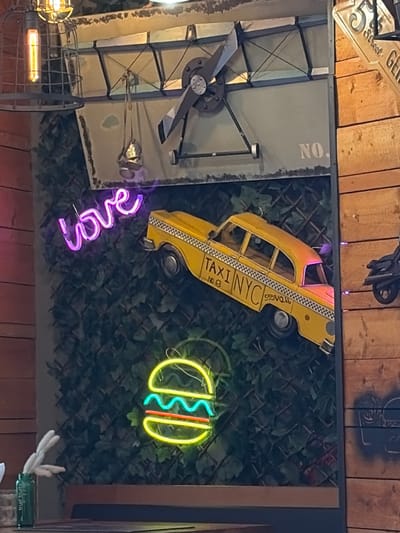 The image captures an eclectic, industrial-themed interior, likely a restaurant or bar, in Essen, Germany. The scene is dominated by decorative elements attached to walls, which are composed of wooden planks, distressed gray panels, and a green, ivy-like covering designed to mimic a hedge.
A prominent yellow model taxi, adorned with "TAXI NYC" and "NO. 8" text on its side, is mounted on the green wall, suggesting a New York City theme. Below it, a vibrant purple neon sign spells out "love" in cursive. To the right of the taxi, another neon sign depicts a stylized hot dog with yellow buns, a green zigzag, and a red line, representing a hot dog with condiments.
Various other decorative objects are visible throughout the space. On the upper left, part of a metallic street-style sign reads "Street Geo." Below it, a large, dark gray propeller-like mechanism is affixed to a distressed gray panel that also displays the letters "NO." vertically. Near the bottom left, an industrial-style light fixture with an orange glow is partially visible within a metal cage. The upper wooden wall also features some dark stenciled graffiti. In the bottom right corner, a partially visible green object shows the inverted text "TRUE FRUITS." The overall ambiance suggests an evening setting or low lighting due to the illuminated neon signs and artificial lights. No people are visible.