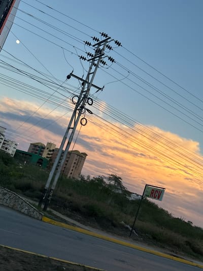 A medium-shot photograph captures a street scene in Pampatar, Venezuela, during what appears to be sunset or sunrise. The sky displays a gradient of blue at the top, transitioning to soft pinks and oranges as it nears the horizon, with scattered clouds adding texture. Dominating the center-left of the frame is a tall, silver utility pole with numerous electrical wires extending from it, creating a complex network across the sky. To the left of the pole, a faint, white moon is visible against the blue sky.
In the background, several multi-story buildings are scattered amongst lush green vegetation. One prominent building, painted green, stands out, with other residential-looking structures visible further to the left and right. The foreground features a paved road with a yellow painted curb. To the right of the utility pole, a signpost with a rectangular sign reading "RCV" in red letters against a green background is planted on a grassy hillside. The scene is tilted slightly, giving a dynamic feel to the composition. No people are visible in the image.