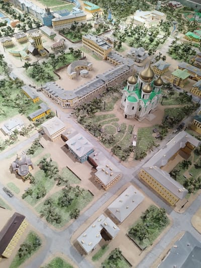 This is a high-angle aerial photograph of a detailed architectural model of a historical city, likely a religious or governmental complex, with the prominent landmark of a large church with golden domes. The model depicts a sprawling urban landscape with numerous buildings of various architectural styles, including classical and baroque influences. These structures are constructed with materials that mimic stone, plaster, and wood, with some featuring painted details in white, yellow, green, and blue.
The scene is populated with miniature trees, shrubs, and landscaping that create a park-like atmosphere interspersed with the urban development. Paved pathways and roads wind through the model, suggesting a functional layout. Small details like benches, streetlights, and even miniature vehicles add to the realism. The model is set in what appears to be a historical city in Pushkin, Russia, given the architectural styles and the characteristic onion domes on the central church.
The lighting in the photograph suggests it was taken indoors under artificial light, as there are no discernible shadows indicative of natural sunlight, and the overall illumination is even. The time of day and weather are not discernible from the model itself. There is no visible text within the image. The overall impression is one of meticulous craftsmanship and a preserved historical environment, likely part of a museum exhibit.
