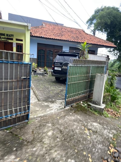 The image shows a view through an open metal gate of a driveway and a house in Lembang, Indonesia (latitude: -6.825, longitude: 107.608). The gate is made of metal bars painted blue and green, and it is partially open. The driveway is paved with gray concrete tiles. A black SUV with a "Jeep" spare tire cover is parked in the driveway. The house is a two-story building with a red tile roof and blue painted walls. The house has windows with white frames, and there is a sign that says "Home Beauty" on the side of the house. The driveway is bordered by a grassy area with some plants growing. The sky is cloudy and gray. There are power lines running overhead.