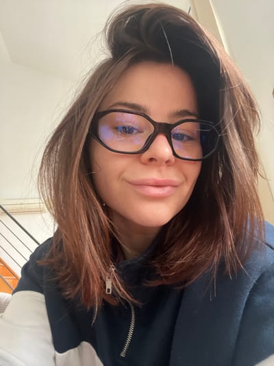 An indoor, close-up, head-and-shoulders portrait depicts a female individual, estimated to be in her twenties or thirties, centered in the frame. She has shoulder-length, medium brown hair, styled with some volume on top, and is looking directly at the camera with a slight smile. She wears dark, octagonal-framed eyeglasses, which display prominent blue light reflections on the lenses. Her lips are closed. She is dressed in a dark blue quarter-zip top with a partially visible white panel on the left upper arm/shoulder area. A silver-colored metal zipper is visible on the front of her garment, pulled partially down. The background to the left shows a white wall, a segment of a black handrail from an ascending staircase, and a white wall-mounted appliance, possibly an air conditioning unit, situated higher on the wall. The background to the right is a plain light-colored wall. The scene is located within the city of Roubaix, France.