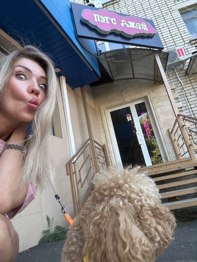A blonde woman with her hair pulled back and a pink shirt is posing for a selfie with her dog in front of a building. The building has a blue awning and a sign that says "ПЭТ С ДЖОЙ" in pink cloud-shaped letters. There is a glass door with a neon sign that says "ГРУМИНГ" in pink letters. The woman has her lips pursed and her eyes wide. The dog is a light brown poodle with curly fur. The scene appears to be in Armavir, Russia.