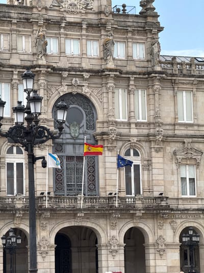 The image shows a section of an ornate stone building with multiple windows and decorative architectural elements. The building's facade is constructed from light-colored stone blocks, featuring carved details, columns, and arched doorways. Several flags are prominently displayed, including the flag of Spain and the flag of the European Union, along with a regional flag that appears to be the flag of Galicia. A street lamp with multiple globes stands in the foreground, adding to the urban setting. The scene suggests a daytime setting with a clear sky. The presence of the flags and the architectural style point to a location in Spain, likely a public building or monument. Visible text on the building reads "CABEZA GVARDA".