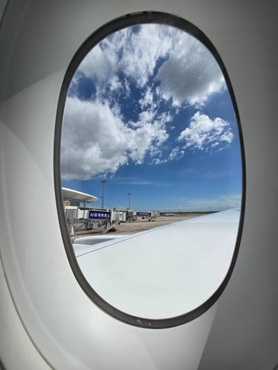 The image is taken from inside an airplane, looking out of a circular window at an airport tarmac on a clear, sunny day. The sky is a vibrant blue with fluffy white clouds scattered across it.
On the tarmac, several jet bridges are connected to a terminal building, with what appear to be a few parked airplanes visible in the distance. Lights and poles are also dotted across the tarmac. The wing of the airplane the photo is taken from is visible on the right side of the frame, extending upwards and slightly to the right. There is some text on a blue banner attached to one of the jet bridges, which appears to be in Chinese characters. The text reads "AI 就用阿里云" which translates to "AI use Alibaba Cloud".
The scene suggests the airplane is either about to depart or has just arrived at the airport. The bright sunlight indicates it is daytime. The clean, blue sky and scattered clouds imply fair weather conditions. The overall impression is one of travel and transit, with the expansive sky and the organized airport infrastructure contributing to a sense of anticipation or arrival. The orientation of the image is tilted, giving a dynamic perspective of the scene.