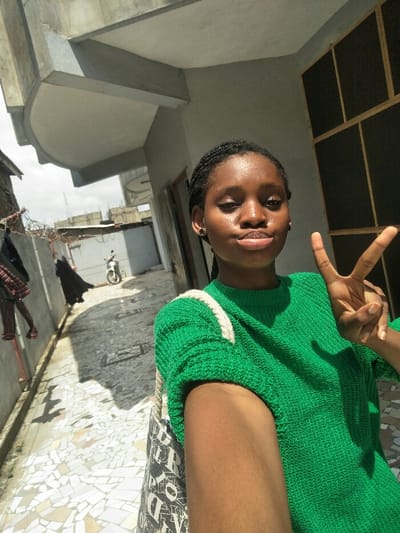 An outdoor daytime scene in Cotonou, Benin, depicts a young female subject captured in a selfie. The female, positioned on the right, wears a green knitted short-sleeved sweater and carries a black and white patterned tote bag with a thick white braided strap over her left shoulder. Her hair is styled in braids, and she has dark stud earrings. She directly faces the camera, her lips pursed, and her left hand is raised, forming a V-sign with the index and middle fingers. Her skin exhibits a noticeable sheen. The background features a narrow concrete pathway with a mosaic-patterned tiled surface. To the left, laundry hangs on a line, and further back, a white scooter is parked. A low wall topped with barbed wire is visible. Above the subject is a concrete architectural overhang, and to her immediate right, a building wall with a dark-paned, grid-patterned window. The sky in the distance appears light and hazy above additional urban structures.
