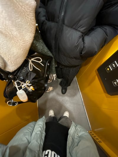 The image displays a top-down view inside an elevator with bright yellow walls and a dark grey floor exhibiting minor speckling. On the right wall, a black control panel features illuminated white up and down arrow buttons, a door-open symbol, and a blue indicator light. Three individuals are visible. In the foreground, the photographer wears a light blue puffer jacket, a black top with partial white lettering, dark trousers, and white sneakers. To their immediate left, a person wears a cream-colored plush garment and a dark green patterned scarf. A black patent leather-like handbag rests between them, adorned with a pearl bow, a black fabric bow, and a keychain holding a white-cased clear bottle, possibly hand sanitizer, along with a gold bee charm. This individual's feet are clad in light brown flat shoes. To the right, another person wears a black puffer jacket, dark denim trousers, and black boots. All individuals are standing. The location is an elevator in Capital City of Prague, Czechia.