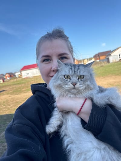 A person is holding a long-haired gray and white cat in their arms. The person is wearing a dark-colored hooded sweatshirt and has fair skin, blonde hair pulled back, and blue eyes. A red string bracelet is visible on their wrist. The cat has green eyes and is looking directly at the camera.
The background includes a blue sky and a grassy field. A few buildings are visible in the distance, including a white building with a red roof. This scene is in Ферма, Russia.