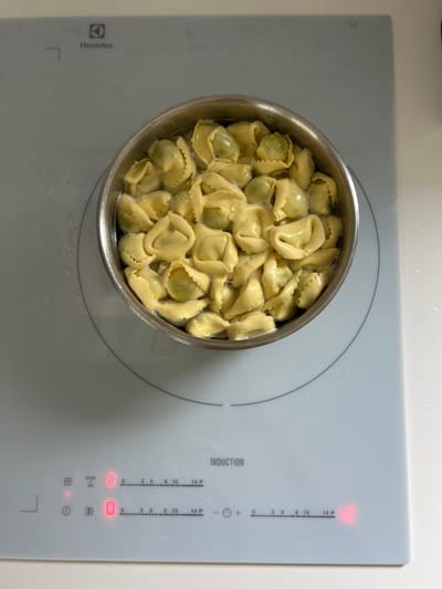 A stainless steel pot filled with cooked tortellini sits on an Electrolux induction cooktop. The pot is positioned on the right side of the cooktop. The cooktop is on, and the timer is set to zero. The timer is blinking, indicating that it is counting down. The tortellini are covered in a clear broth. The cooktop is white with gray markings. The background is white. The location is a kitchen in Balaruc-les-Bains, France.