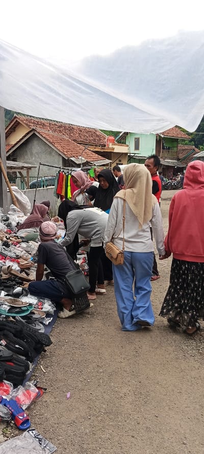 A group of people are gathered in front of a stall in the town of Kutawaringin, Indonesia, located at coordinates -6.974 latitude, 107.519 longitude. The stall is covered by a white tarp. The people are looking at and possibly purchasing shoes and sandals that are displayed on the ground. Several bags and other items are scattered around the stall. The people are dressed in a variety of clothing, including brightly colored shirts, long pants, and head scarves. Some are wearing backpacks. There are several houses and other buildings in the background, including a large green building with a balcony.