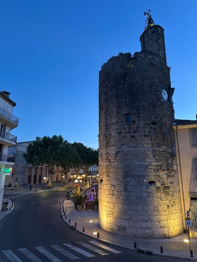 The image shows a view of the Tour de l'Horloge in Anduze, France, at dusk. The stone tower, illuminated by spotlights, stands prominently on the right side of the frame. A clock face is visible on the tower. In the background, a street curves to the left, lined with buildings and trees. Pedestrians and a baby stroller are visible on the sidewalk. The sky is a deep blue, indicating the transition from day to night.