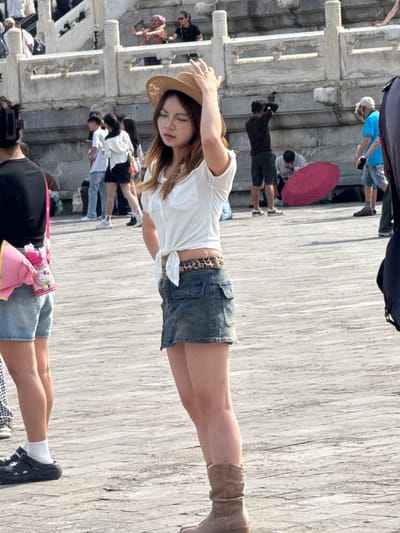 In Dongcheng District, China, a young woman wearing a straw hat, white short-sleeved shirt tied at the waist, and a denim mini-skirt stands on a paved walkway. She is facing sideways with her head tilted back and her eyes closed. Her right hand is raised and touching her hat. She is wearing tan ankle boots. To her left, another person wearing a black top and denim shorts is visible from the waist down. A pink Hello Kitty water bottle is clipped to their belt. In the background, several other people are walking and standing around, with stone architecture and steps visible. A bright red umbrella lies on the ground near some of the background figures. The scene appears to be a public outdoor space with historical architecture.