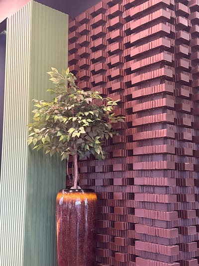 This image depicts an indoor architectural scene, presumed to be located within the city of Prokhladnoe, Russia. The environment is devoid of any individuals, actions, or interactions. A prominent feature on the left side of the frame is a tall, artificial plant, characterized by numerous green leaves and visible roots, standing in a large, cylindrical pot. The pot exhibits a glossy finish, transitioning from a dark, speckled brown at its base to a vibrant amber and orange rim at the top.

The background comprises two distinct wall treatments. To the left of the plant, a vertical structure is visible, composed of light green panels with a uniform, vertical ribbed or fluted texture. Further to the left, a sliver of another, lighter-colored ribbed panel is discernible. Dominating the right side of the image is a striking wall surface constructed from reddish-brown, rectangular blocks. These blocks are arranged in an intricate, three-dimensional pattern, creating a stepped and recessed effect that imparts a strong geometric and textural quality to the wall. The lighting appears consistent with an indoor setting, casting subtle shadows that enhance the depth and patterns of the architectural elements. The overall impression is that of a meticulously designed interior, possibly a commercial or public space.