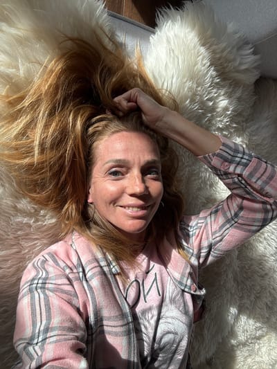 A woman with blonde hair lies on a white fluffy rug. She is wearing a pink plaid shirt. She has her right hand raised, resting on her head. The woman is smiling at the camera. Sunlight is casting shadows on her face and the rug. The background appears to be a wooden floor and a couch. There is no identifiable information indicating the location is Steklyannyi, Russia.