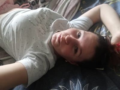 A woman is lying on her back on a bed, facing the camera. She is wearing a light gray t-shirt with small black polka dots and a pattern of clear beads or sequins on the upper chest area. Her right arm is extended above her head, and her left arm is bent at the elbow, resting near her head. The background consists of a dark blue patterned bedspread and other bedding materials, including a pink fabric and a leopard-print blanket, visible in the upper left corner. The lighting is dim, and the image is taken from a close-up perspective, focusing on the woman's face and upper body. There is no indication of the specific location within Zlatoust, Russia.