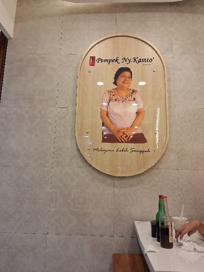 A light wood sign hangs on a wall with light-colored tiles. The sign is oval-shaped with a woman’s portrait, text reading “Pempek Ny. Kamto” and a line in Indonesian. At the top right, a person with a white cloth wipes a table. A bottle with a green neck and brown body sits on the table. A wooden object is under the edge of the table.