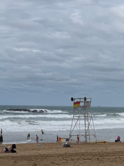 This is a photograph taken at the beach in Biarritz, France, on an overcast day. The scene depicts a sandy beach with people engaged in various activities. In the foreground, several individuals are relaxing on towels and blankets, some appearing to read or rest. Further down the beach, closer to the water, a group of people are in or near the ocean. Some are standing in the shallow water, while others are on surfboards, suggesting surfing or paddleboarding. A lifeguard tower stands prominently on the right side of the beach, a common fixture on such shorelines. In the background, waves crash against the shore, indicating moderate surf conditions. The sea extends to the horizon, where a hazy sky with clouds dominates the upper portion of the image. The overall atmosphere is one of a typical beach day, albeit under cloudy skies.