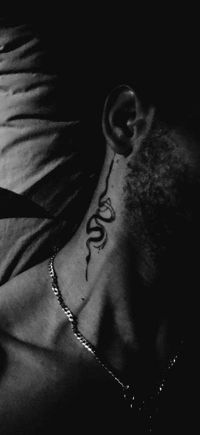 A close-up, grayscale, low-light image shows a person's neck and upper shoulder area, focusing on a snake tattoo. The tattoo is located on the left side of the neck, extending slightly upwards towards the ear. A silver chain necklace is visible around the neck, resting on the collarbone. The background consists of dark, textured fabric, possibly bedding or clothing. The lighting is dramatic, casting deep shadows and highlighting the skin texture and the tattoo. The person's ear is partially visible at the top right of the frame. The image does not contain any information related to Kostenets, Bulgaria.