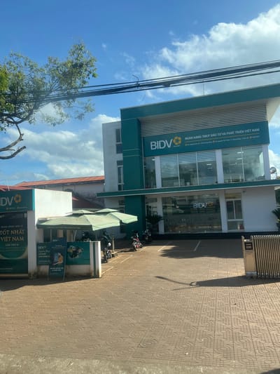 This image depicts the BIDV bank branch located at the Nguyen Tat Thanh x Krong Nang intersection in Krông Năng, Krông Năng District, Vietnam. The main structure is a two-story building with a white facade accented by teal green panels, featuring prominent "BIDV" branding and the full bank name in Vietnamese. Its ground floor consists primarily of large glass windows. To the left, a single-story annex displays additional BIDV signage, including promotional text for "Best Retail Bank in Vietnam." In front of this annex, a large green multi-canopy umbrella shades an area where several motorbikes are parked. Additional motorbikes are visible in the foreground's paved parking lot, which exhibits a distinctive geometric tile pattern. A silver accordion-style gate is partially open on the right side of the parking area. Overhead utility lines and a tree branch with green foliage are visible in the upper left, set against a blue sky with scattered white clouds. The scene is illuminated by strong daylight, casting shadows on the paved surface. No individuals are present in the frame.