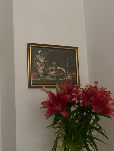 The image displays an interior corner of a room located in Heidelberg, Germany. A rectangular still life painting, framed in gold with a dark inner border, hangs on the textured white wall. The painting features a dark background and depicts a vase of various flowers on the left, a central bowl filled with nuts and dried fruits topped by an ornate gold vessel, a tall glass containing red liquid, a glass pitcher, and several pretzels arranged on a surface. Below and to the right of the painting, a substantial bouquet of vibrant red lilies with green stems and leaves is prominently positioned, partially obscuring the lower wall and extending towards the foreground. No individuals or specific actions are visible.