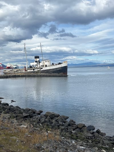 The scene depicts the grounded shipwreck *Desdemona* located in the bay of Ushuaia, Argentina. The ship, an old cargo vessel, is listing to its port side and is situated within a perimeter of large dark grey rocks along the shoreline. Its hull presents a black lower section and a heavily rusted white upper section. A prominent black and white smokestack is visible on the deck, flanked by two unrigged masts. The immediate foreground features a rocky embankment comprised of dark, angular stones, sparse dry vegetation, and scattered debris. The bay's water is calm and grey-blue, reflecting the overcast sky with minimal ripples. In the mid-ground, a small white floating marker or buoy is visible further offshore to the right. The background on the left reveals several active vessels, including a large red and white ship, docked along the distant port area. A distant, hazy blue-grey mountain range defines the horizon beneath a heavily clouded sky featuring some patches of lighter blue. No individuals are present, and no overt actions or interactions are occurring.