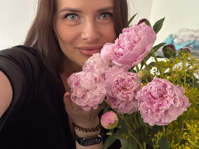 An upside-down close-up portrait features a woman holding a bouquet of flowers, suggesting a selfie taken with the camera inverted. The woman's face, with light-colored eyes and a neutral expression, occupies the lower portion of the frame. Her dark brown hair cascades around her face.
She holds a vibrant bouquet dominated by large, light pink peonies in various stages of bloom, along with unopened buds. Sprigs of yellow, small-flowered foliage are interspersed among the peonies. Part of the woman's right arm is visible, adorned with a black smartwatch on her wrist and a dark, spiral-shaped hair tie or bracelet further up. A simple ring is worn on her right ring finger. She appears to be wearing a dark, possibly black, garment.
The background is predominantly a plain, light-colored surface, likely a wall. Some indistinct, light-toned objects are blurred in the top left corner. The image depicts an indoor setting, and the activity is a self-portrait with flowers. No specific time of day or weather is discernible, and no text is visible within the image. The location context for this media is Rusalevka, Russia.