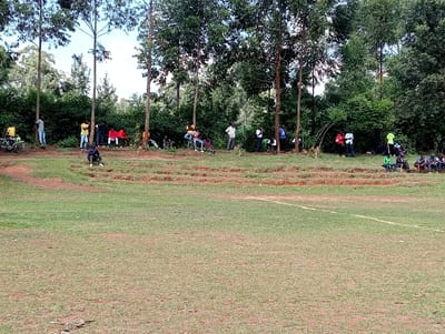 The image shows a grassy field with a dirt track surrounding it. There are a few sets of bleachers in the dirt track. Twenty-two people are standing and sitting in the dirt track, the bleachers, or the edge of the grassy field. There are trees in the background. The scene is located at the Kipkarren High School, in Kipkarren, Kenya. The school is located at 0.601° N, 34.939° E.  There are two bicycles leaning against a tree in the dirt track. Two people are riding the bicycles towards the bleachers. The people in the bleachers are looking down at the field. The people in the dirt track are looking toward the camera. The people are wearing casual clothing.  A bicycle is parked in the dirt track to the left of the bleachers.  The grassy field is a bright, vibrant green. The ground is a dull brown with a slight red tint in the dirt track. The trees are mostly a deep green. There is a line of trees along the edge of the field. The trees are all different heights and sizes.  The sky is a light blue.  The air is clear. The people are enjoying the day. The weather is pleasant.  The image shows a day at the Kipkarren High School in Kipkarren, Kenya.  The image was taken on a bright, sunny day with a clear sky.