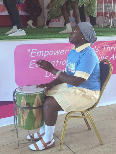 This media file captures a vibrant scene from Kakuri Gwari, Nigeria, likely during a cultural or educational event. The central subject is a person dressed in a blue t-shirt with yellow trim and a light yellow pleated skirt. They are seated on a golden chair and actively playing a drum. The drum is cylindrical, predominantly green with lighter green and wood-toned accents, and secured with metal straps. The person is wearing a grey headscarf, white socks, and brown sandals.

In the background, a large pink banner with white text is visible, partially obscured by the seated individual and others. Text on the banner includes phrases like "Empoweries Thro" and "Girls Through ini," suggesting an event focused on empowerment and education. Other individuals can be seen in the background, some wearing green attire with intricate patterns, and others in dark trousers. The setting appears to be outdoors or in a large, open indoor space with a tiled floor and what looks like artificial grass in the foreground. The lighting suggests it is daytime. The overall mood is one of active participation and celebration, as indicated by the drumming and the colorful attire.