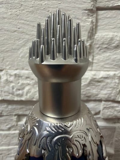 This is a close-up, eye-level, indoor photograph of the metallic cap of a bottle, set against a white brick wall. The cap features a decorative, silver-colored floral motif with intricate detailing. Atop the cap, there is a cluster of numerous small, pointed silver spikes arranged in a radial pattern. The lighting is direct, creating highlights and shadows on the metallic surfaces. There are no people or animals present, and the background is a textured white brick wall. The scene appears to be a still life composition. The photograph does not contain any elements that would indicate its location in Haskovo, Bulgaria.