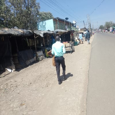 The scene depicts a sunny daytime street in Nkaimurunya ward, Kenya. A paved road occupies the right side of the frame, bordered by an unpaved dirt and gravel shoulder on the left. A male subject, viewed from behind, is walking from left to right on the dirt shoulder. He is wearing a light blue long-sleeved collared shirt, dark trousers, and dark shoes, with a tan or light brown jacket draped over his left arm. To the immediate left of the shoulder, a row of approximately five makeshift market stalls is visible. These stalls are constructed with wooden frames and covered predominantly with black tarpaulin or plastic sheeting. One stall near the foreground contains white rectangular objects, possibly polystyrene boxes. Further down this row, a metal handcart loaded with produce is positioned. Behind the stalls, a multi-story building constructed with light blue corrugated metal siding and featuring balconies is present. A satellite dish is mounted on the roof of this building. Overhead, multiple utility lines traverse the clear blue sky, supported by poles visible further down the road. In the mid-ground and background, multiple pedestrians are observable walking along the road and its adjacent areas.