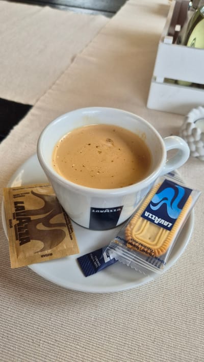 A white ceramic cup of coffee sits on a white saucer, placed on a beige checkered tablecloth. To the left of the cup is a small packet of Lavazza sugar. To the right of the cup is a packaged Lavazza biscuit. The background shows a white decorative wooden box with bottles inside. The scene is indoors. The exact location within Ravda, Bulgaria, cannot be determined from the image.