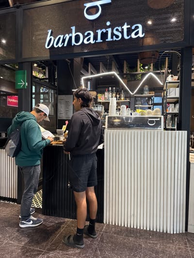 Two men stand at a counter inside a "barbarista" cafe. The first man wears a black jacket, shorts, and black socks with sandals. He looks down at something on the counter. The second man wears a white baseball cap, a green jacket, gray jeans, and blue sneakers, and reads a menu. Behind them, shelves display various items and a zigzag neon light fixture decorates the wall. The cafe's name is displayed prominently in neon letters.