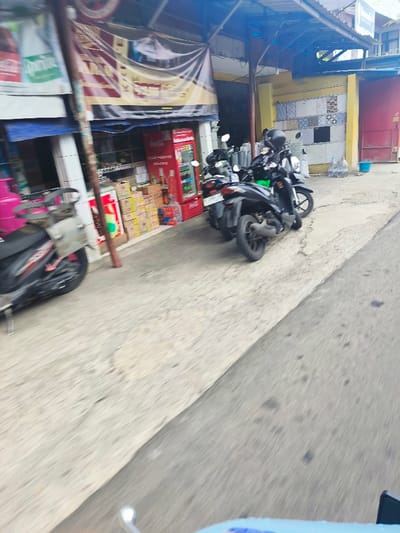 The image shows a scene in Lembang, Indonesia, taken from the perspective of a person riding a motorcycle. Two parked motorcycles are visible on the right side of the image, in front of a small shop with a red Coca-Cola vending machine. The shop appears to be selling various goods, including boxes of bottled drinks and other items. The motorcycle rider is looking ahead toward the road, which extends into the distance. The road is paved and appears to be in good condition. The sidewalk is also paved and is relatively clean. The overall environment is typical of a small town in Indonesia, with a mix of commercial and residential buildings. The image captures a sense of normalcy and everyday life in the area. The time of day is likely daytime.