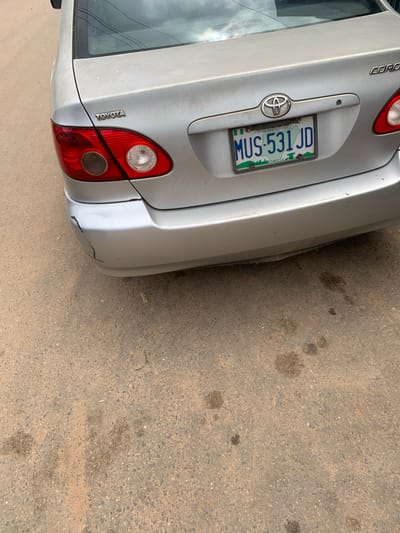 The image shows the rear of a silver Toyota Corolla car parked on a dirt road. The car has a Nigerian license plate that reads "MUS-531 JD". The car has some damage to the rear bumper. The environment appears to be in a dusty, urban area. Based on the license plate and the style of the car, the image is likely taken in Nigeria. Given that the license plate is from Nigeria and the car is a Toyota Corolla, a common vehicle in Nigeria, and considering the context provided, the scene is likely located in Shagamu, Nigeria.