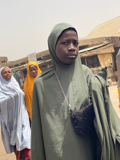 The image depicts three individuals in an outdoor, arid urban environment in Unguwar Gabas, Nigeria, consistent with a market or street scene.
The central figure, a young person with dark skin, stands in the foreground, directly facing the camera with a neutral expression. They wear a dark olive-green full-length garment with an integrated head covering. A small, black, fluffy faux fur bag with a silver chain strap hangs from their left shoulder.
Behind and to the left of the central figure is an adult female with dark skin, wearing a light blue head covering and garment, with a visible patterned skirt beneath. Her mouth is slightly open, and she looks to the viewer's left.
Behind the central figure, slightly to the left, is another adult female with dark skin, wearing a bright mustard-yellow head covering and garment. She looks towards the viewer with a focused expression.
The background consists of several single-story, rudimentary structures, likely market stalls or shops, with corrugated metal roofs, visible wooden supports, and walls constructed from local materials. Some structures feature light-colored fabric awnings. The sky is bright and hazy, indicating strong daylight. The ground appears unpaved.
