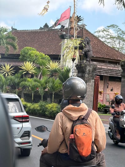 A medium shot captures a person riding a motorcycle in Tampaksiring, Indonesia. The rider is wearing a dark grey helmet and a light brown hooded jacket over dark pants. A dark backpack with an orange pouch featuring the Eiger logo and a water bottle is strapped to their back. To the left, the rear of a silver car is visible. To the right, another person on a scooter wears a black helmet and a light brown shirt. Behind the motorcyclists, a building with a red tile roof and ornate stonework is prominent, adorned with tropical plants and a flagpole flying a red and white flag. The scene is set against a backdrop of lush greenery and a cloudy sky.