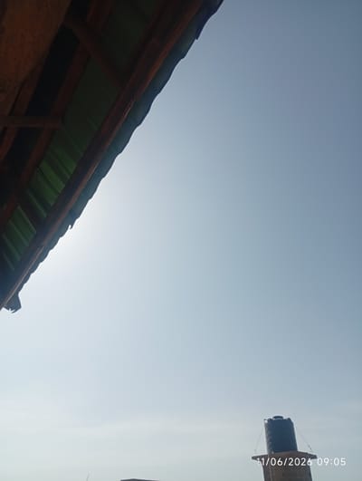 The image, taken on June 11, 2026, at 09:05, shows a bright, clear sky above what appears to be a rooftop in DU, Nigeria. In the foreground, a portion of a wooden roof structure with a green corrugated roof is visible at an angle from the left side of the frame. Towards the bottom right of the image, a black water tank is perched on a raised platform, supported by thin wires. The water tank is partially obscured by a brick structure below it. The sky is a uniform light blue, with the sun appearing to be somewhere behind the roof structure, causing some glare. No people are visible in the image.