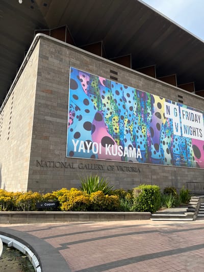 This image captures the exterior of a large building, identified as the National Gallery of Victoria, located in Southbank, Australia. The building features a facade constructed from light grey, rectangular stone or concrete blocks.
Dominating the building's upper section is a very large, colorful banner promoting an exhibition or event. The banner prominently features the name "YAYOI KUSAMA" in large white capital letters. Above this, the text "NG FRIDAY NIGHTS" is visible. The background of the banner is an abstract, vibrant composition of undulating forms in shades of blue, green, purple, and pink, all covered with numerous black polka dots of varying sizes.
Below the banner, carved into or raised from the building's facade, are the words "NATIONAL GALLERY OF VICTORIA" in a classic serif font. To the left of the main building, a modern, angular roof structure with dark gray paneling is visible against a clear, light blue sky.
Along the base of the building, a strip of dense landscaping features various green and yellow bushes, shrubs, and a palm-like plant. Adjacent to the landscaping is a paved area, likely a sidewalk or plaza, made of reddish-brown pavers arranged in a linear pattern with darker stripes. A small black sign with a white arrow pointing left and the word "Carpark" is partially visible within the landscaping.
The scene is bathed in bright daylight, indicating a sunny day. No people are visible, and there is no specific activity or event taking place other than the advertisement for the art exhibition. The overall impression is one of a contemporary urban cultural institution.