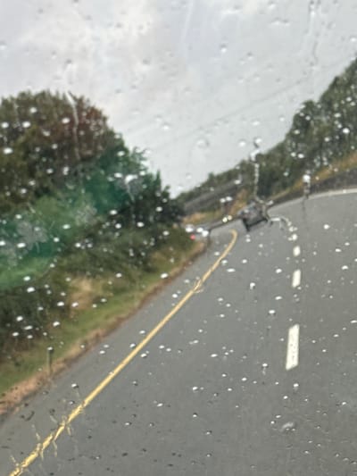 The image is a photograph taken from inside a vehicle, looking through a rain-streaked windshield. The scene depicts a wet road with a yellow dividing line, bordered by grassy verges and trees. In the distance, on the road, several vehicles are visible, including what appears to be a dark-colored car. The sky is overcast and gray, indicating a rainy day. The specific location in the city of Gorey, Ireland cannot be determined from the visual information provided.