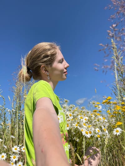 In the city of Сям-Можга, Russia, a young woman with blonde hair pulled into a ponytail, wearing a bright green t-shirt and earrings, is captured in a selfie. She is surrounded by a field of white and yellow wildflowers under a clear blue sky. The woman is shown from the shoulders up, facing to the right with her eyes closed and a slight smile. Her left arm is extended towards the camera, holding a bouquet of the wildflowers she is amidst. The background is dominated by the vibrant blue sky and the lush green and yellow hues of the meadow, with some taller grasses framing the scene.