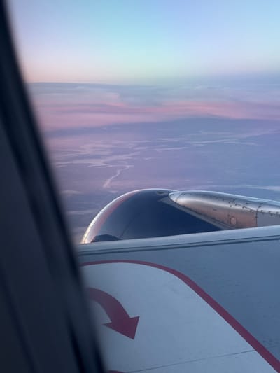 An aerial view from an airplane window captures a landscape beneath a soft, gradient sky transitioning from pale blue to pink and lavender hues. The ground below appears to be a network of rivers or waterways winding through a muted, possibly arid terrain. In the foreground, a portion of the airplane's wing is visible, featuring a metallic engine nacelle with visible rivets and panel lines. The wing also displays a red curved arrow symbol, indicating an emergency exit. The window frame, a dark, blurred element, partially obscures the view. The scene suggests a flight over a remote or undeveloped area, possibly at sunrise or sunset given the atmospheric colors. There is no visible indication of the city of Abakan, Russia.