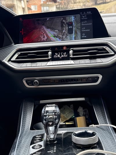 The image shows the interior of a car, most likely a BMW based on the design of the dashboard, infotainment system, and gear selector. The infotainment screen is displaying a camera view labeled "Front," with menu options visible, including "Park Assist," "Reversing Assistant," "Camera picture," and "Settings." The climate control panel indicates a temperature of 20.0°C (68°F). The gear selector is a crystal-style knob with a "P" (Park) indicator. The center console includes buttons for features like "Auto Start/Stop," parking sensors, and a "Sport" mode. The interior trim features a patterned design. The image does not show the location in the city of Lilkovo, Bulgaria.