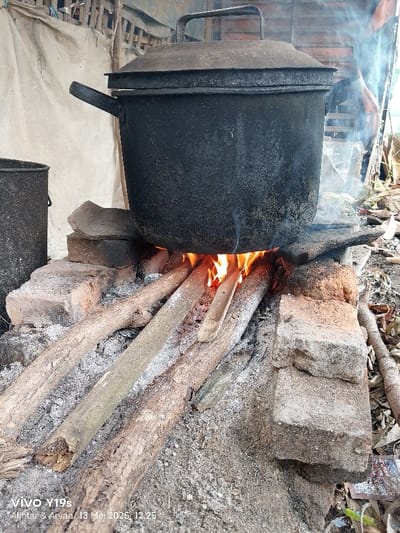 A large, black metal pot with a lid sits atop a small fire made of four wooden logs on a dirt and brick base. The pot has a handle on the left side. The fire is small and contained, with flames visible.  The pot is sitting on four pieces of wood with a fire burning beneath it.  The scene is taken outside, most likely in the backyard of a home. The scene is located at the coordinates -6.419, 107.978, which corresponds to a rural area in the city of Anjatan, Indonesia.  There are no people visible in the image. The pot appears to be used for cooking. The image was taken on a phone, as evidenced by the watermark on the bottom right corner.